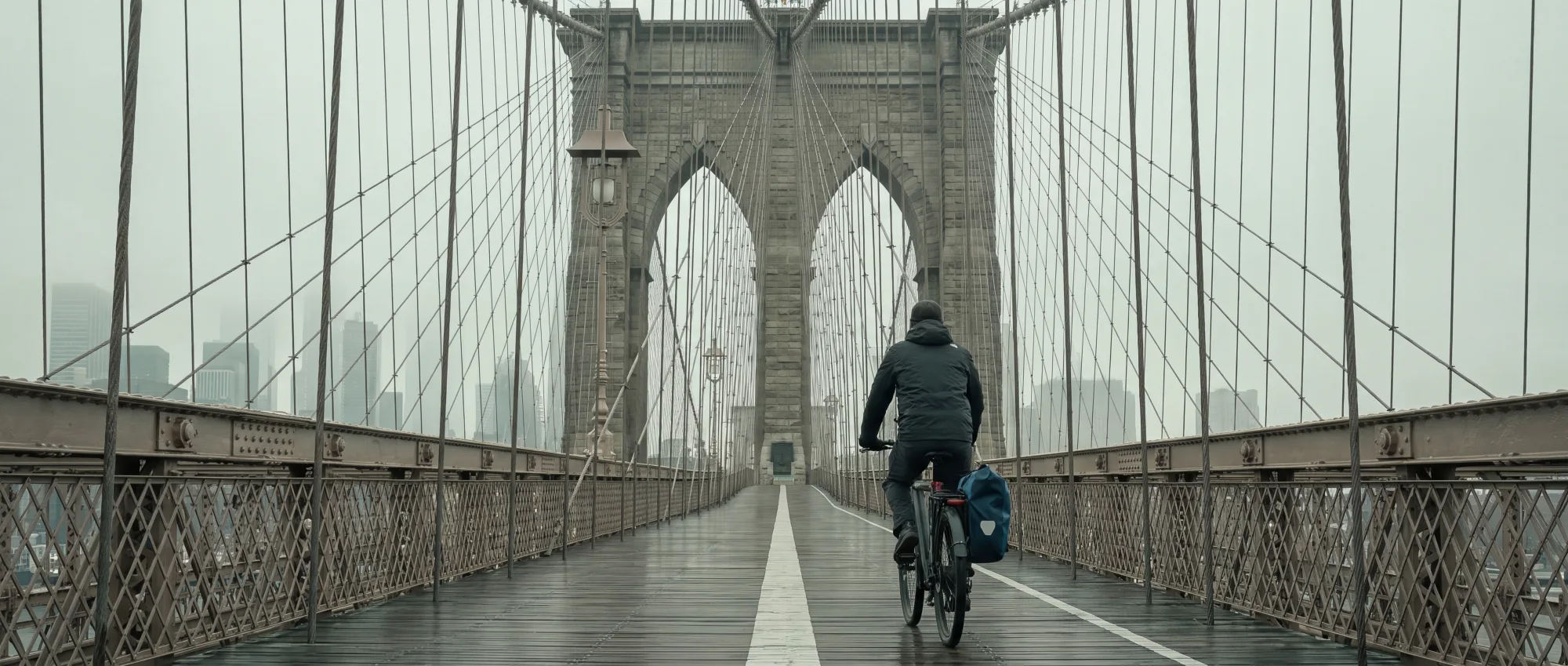 A lone commuter on an e-bike crossing the Brooklyn Bridge walkway with the Manhattan skyline behind