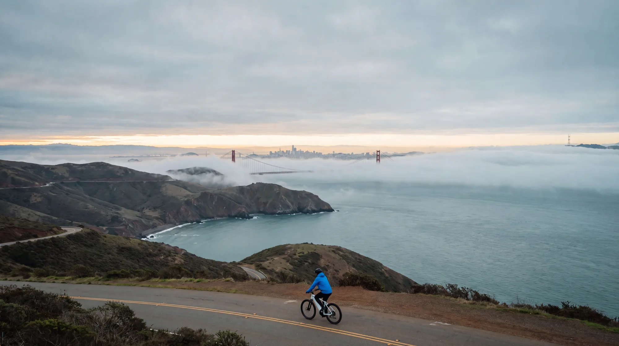 A lone cyclist on a coastal headland road with the Golden Gate Bridge and the San Francisco skyline in fog behind